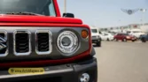 2026 Suzuki Jimny GLX AT frontend closeup showcasing the distinctive round LED headlight, chrome-accented grille, and red body paint