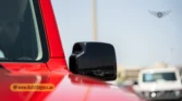 2026 Suzuki Jimny GLX AT side profile view showing the black side mirror cap, red body panel, and watermarked AutoMagnus.ae logo